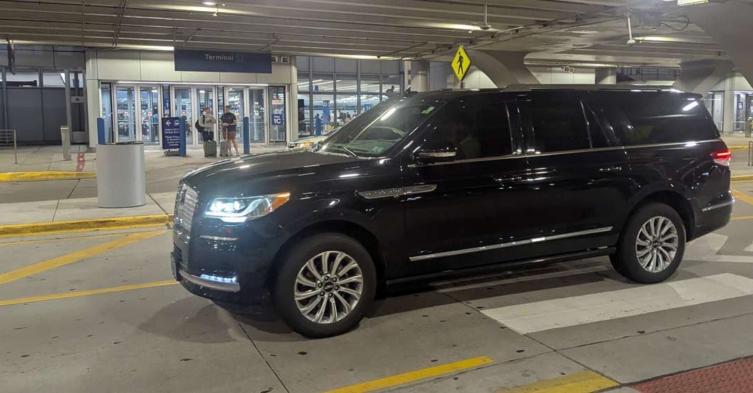 Black SUV/van-like vehicle parked at airport curb with pedestrians in the background near the terminal entrance at night.