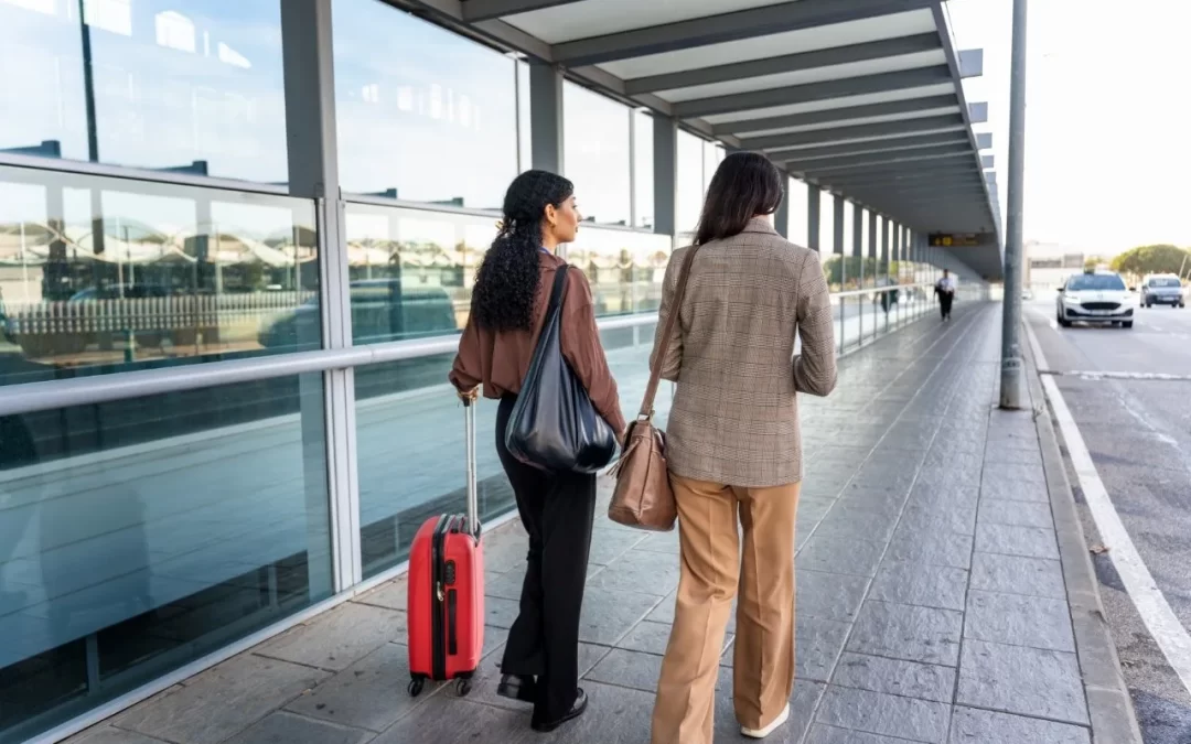 Two women walking beside glass terminal walls with a red rolling suitcase