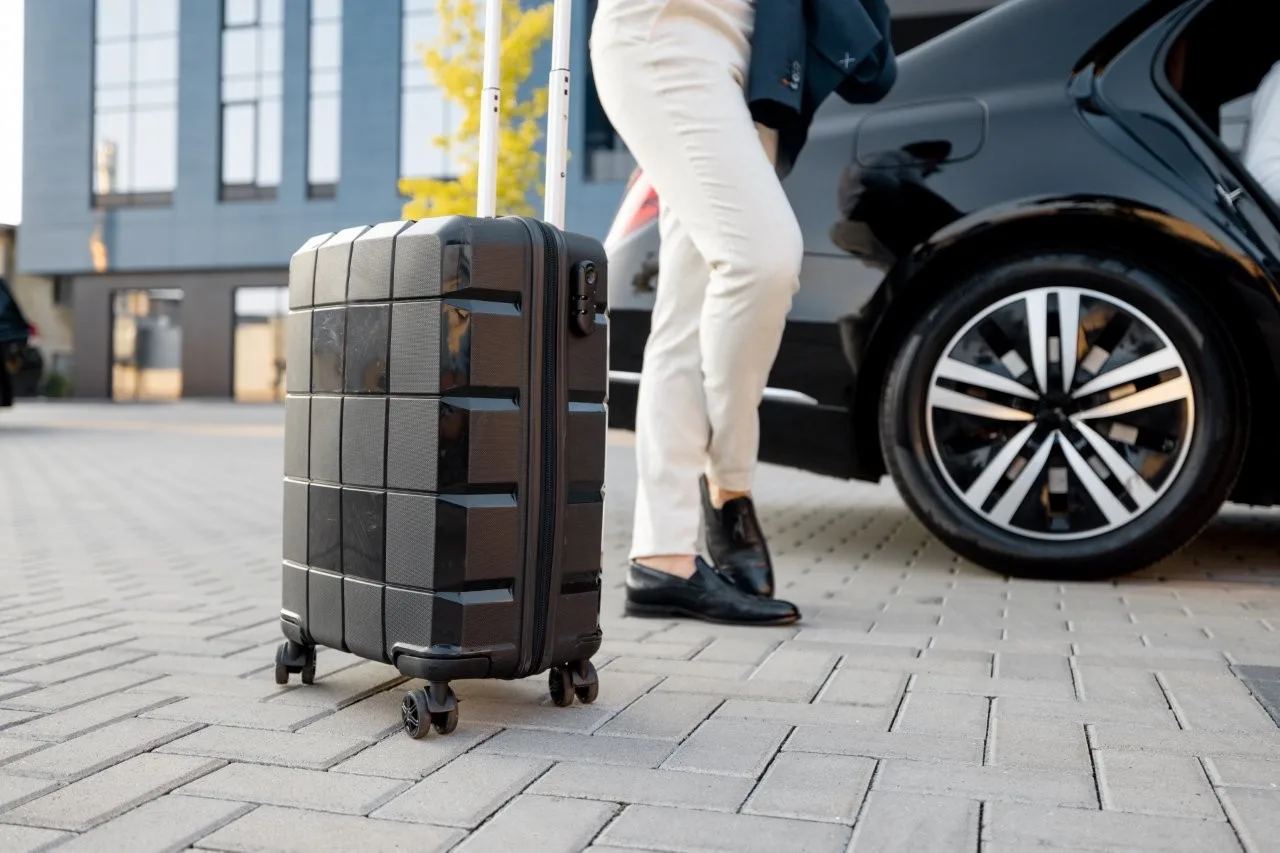 A rolling suitcase beside a traveler in light trousers and a black car outside a building