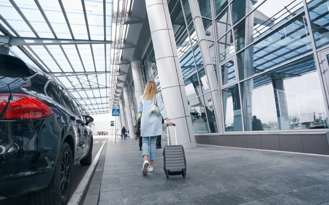 A woman in a light blue suit pulling a gray suitcase beside a black car and glass terminal