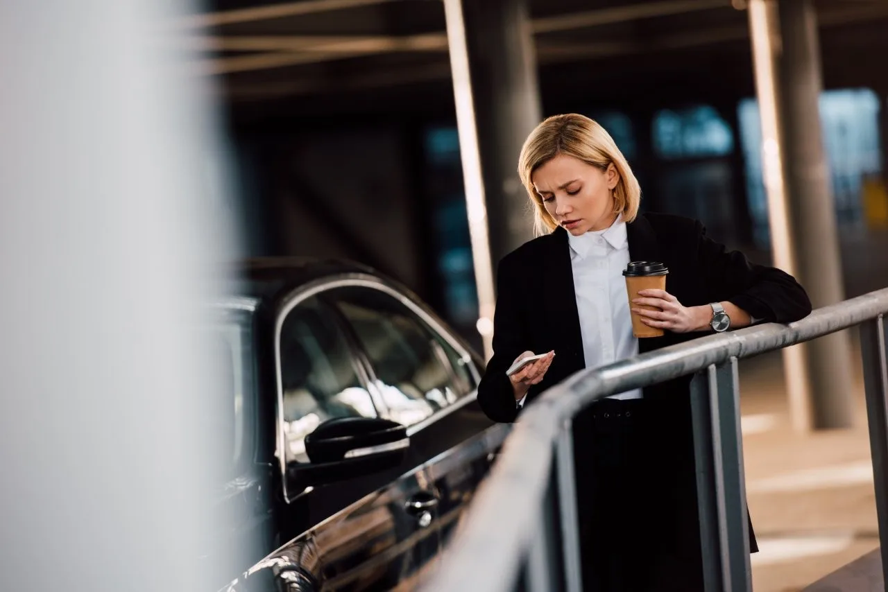 A woman in a black suit holding a phone and coffee beside a black car and metal railing