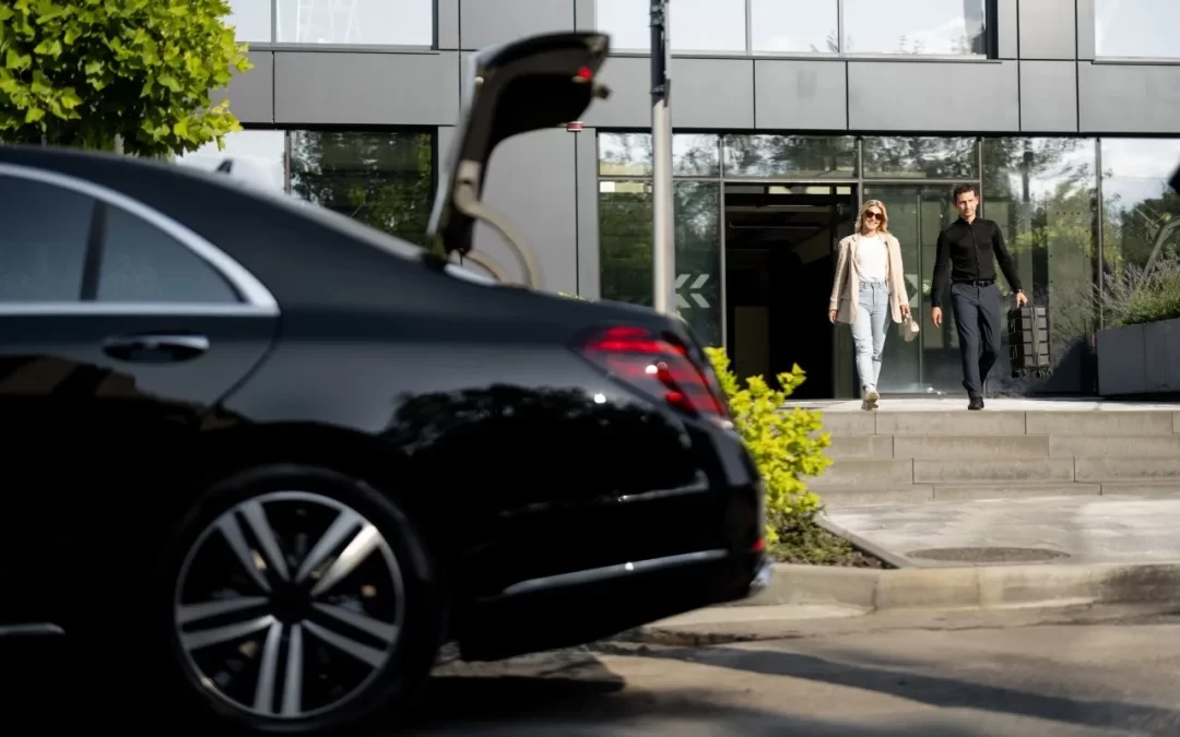 Two business travelers outside a glass-fronted building beside a black sedan with an open trunk
