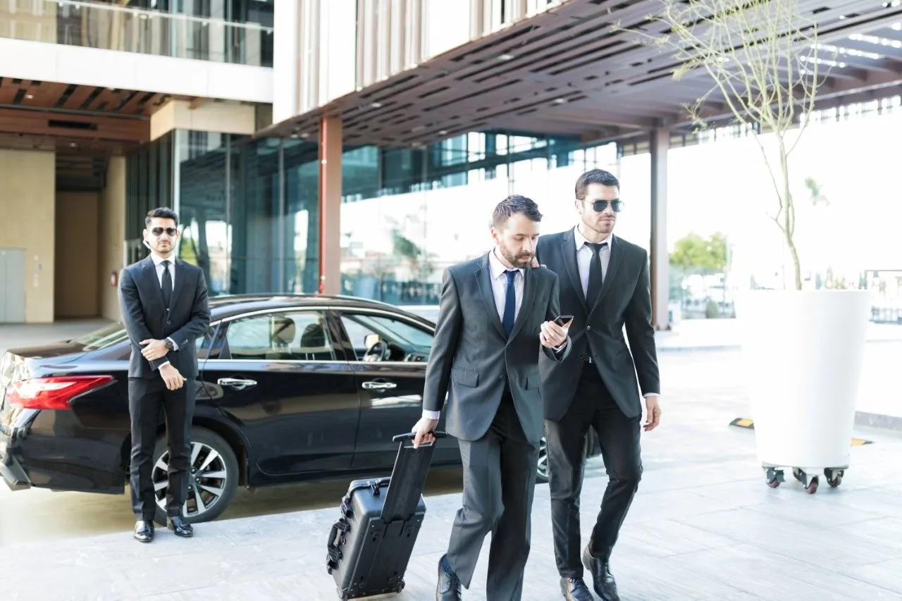 Three men in dark suits beside a black sedan, one pulling a suitcase