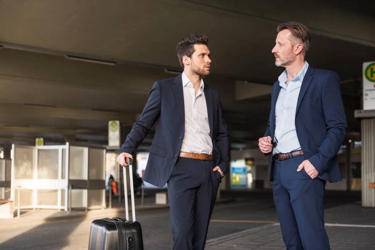 Two men in suits talking under a covered outdoor area beside a suitcase
